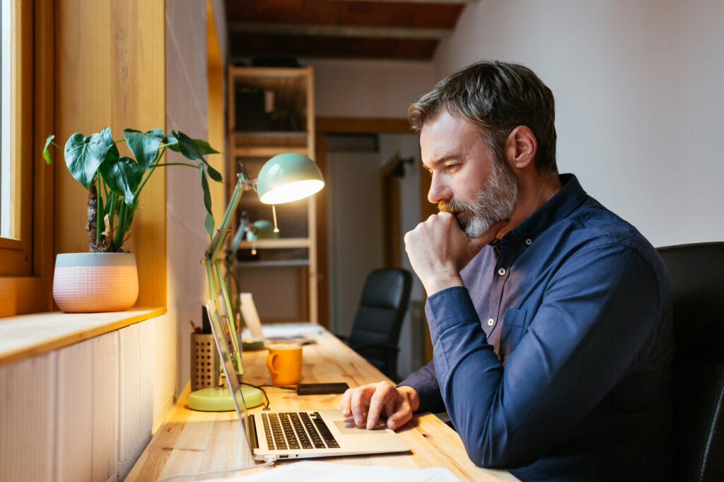 Side view of smart bearded man rubbing chin and thinking while sitting at desk and using laptop in cozy workspace at home