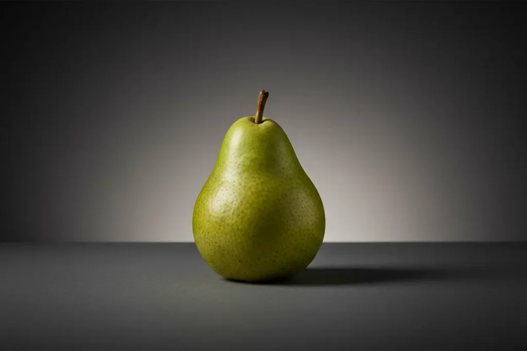 Single green pear on a dark-to-light gray background, shaped by soft light and deep shadows to highlight texture and form.